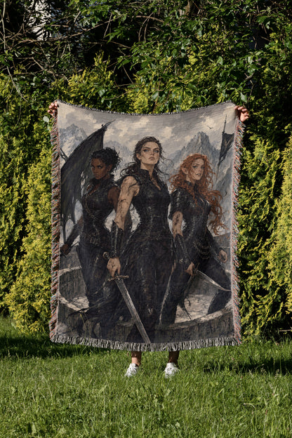 A person is holding up a large tapestry or blanket featuring three women in dark clothing, each holding a sword, in a grassy outdoor setting with trees in the background.