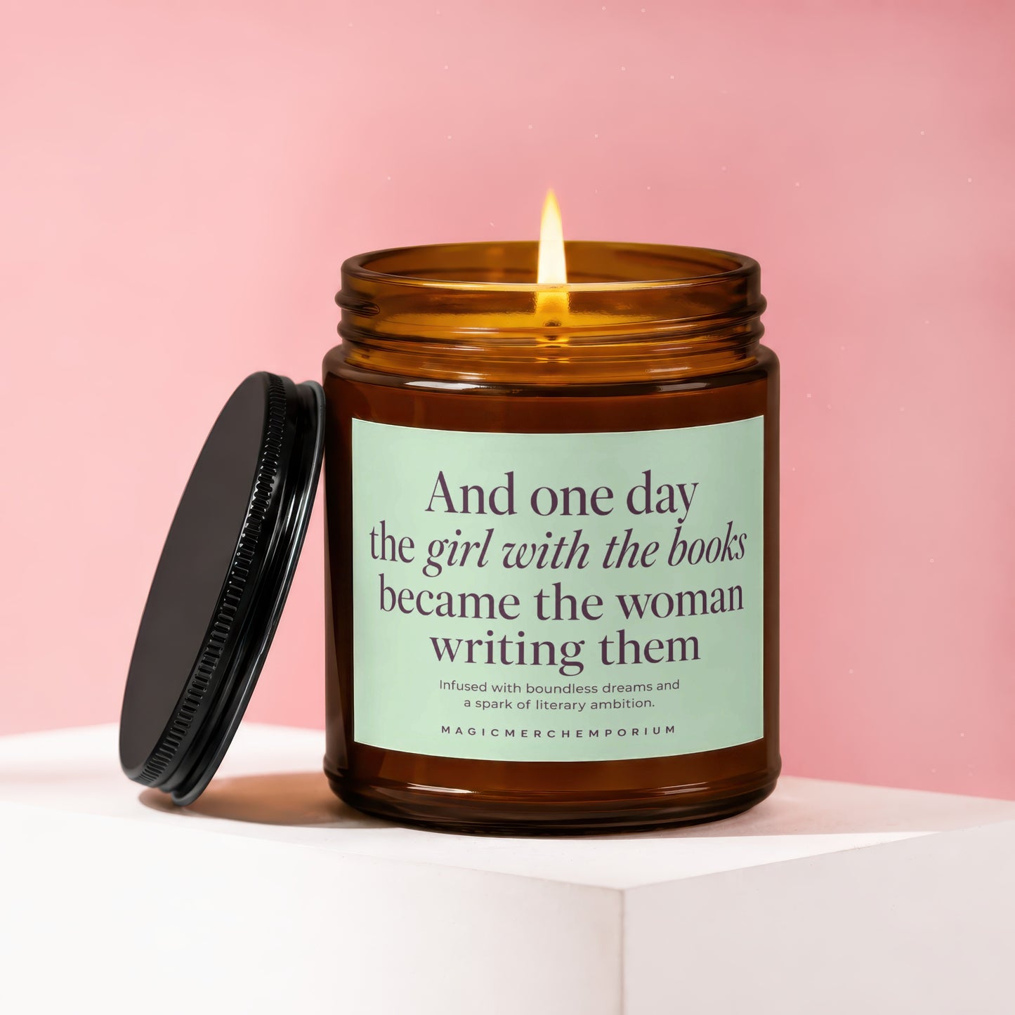 Amber candle with lit wick, lid beside it, reads "the girl with the books became the woman writing them."