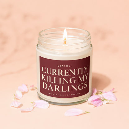Glass jar candle with burning wick and dark red label reading "STATUS: CURRENTLY KILLING MY DARLINGS".