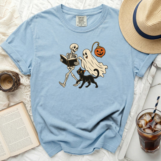 Skeleton reading book, ghost with pumpkin, and black cat illustration on blue t-shirt.
