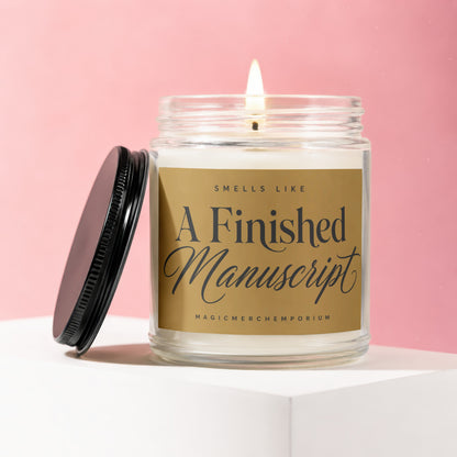 A lit candle in a glass jar with a gold label that says "SMELLS LIKE A FINISHED MANUSCRIPT".