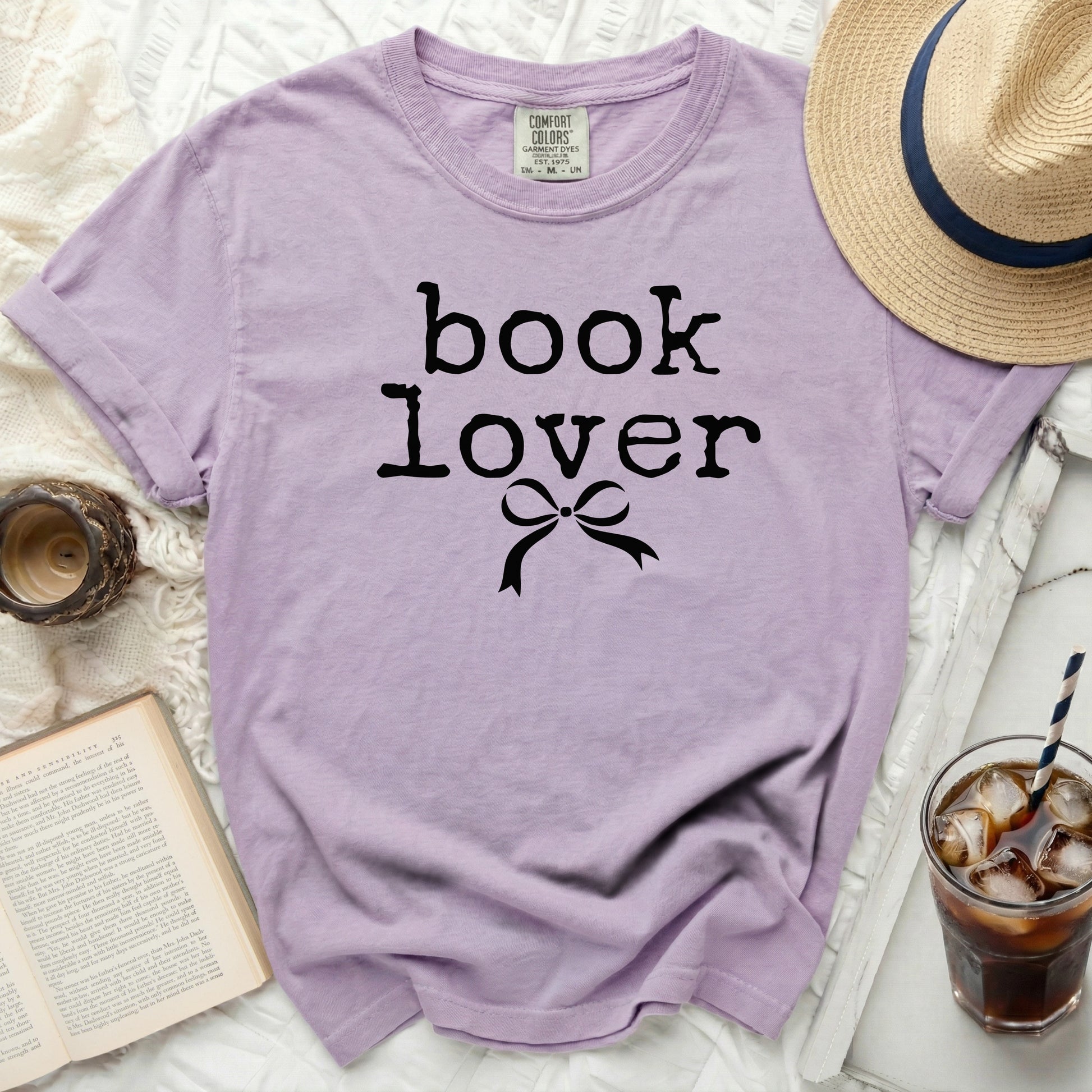 Purple t-shirt with "book lover" graphic and a bow, next to a book and drink.
