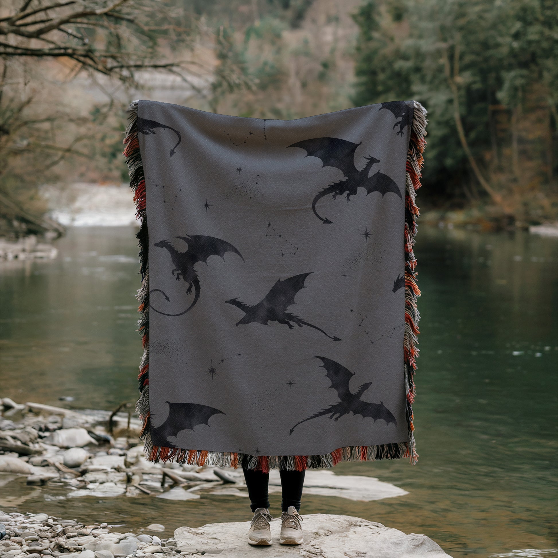 A person is standing on a rock, holding a large blanket with a dragon design on it, with a body of water and trees in the background.
