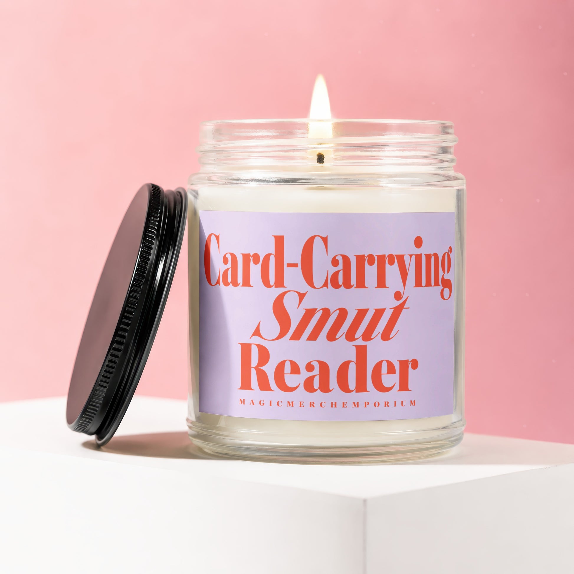 Candle with 'Card-Carrying Smut Reader' text on lavender label, lid off, on white cube.