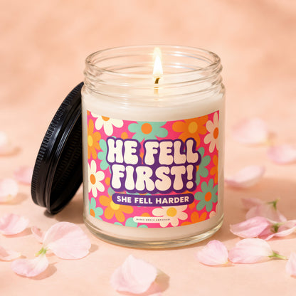 Candle with lit flame and "HE FELL FIRST! SHE FELL HARDER" text on floral label.
