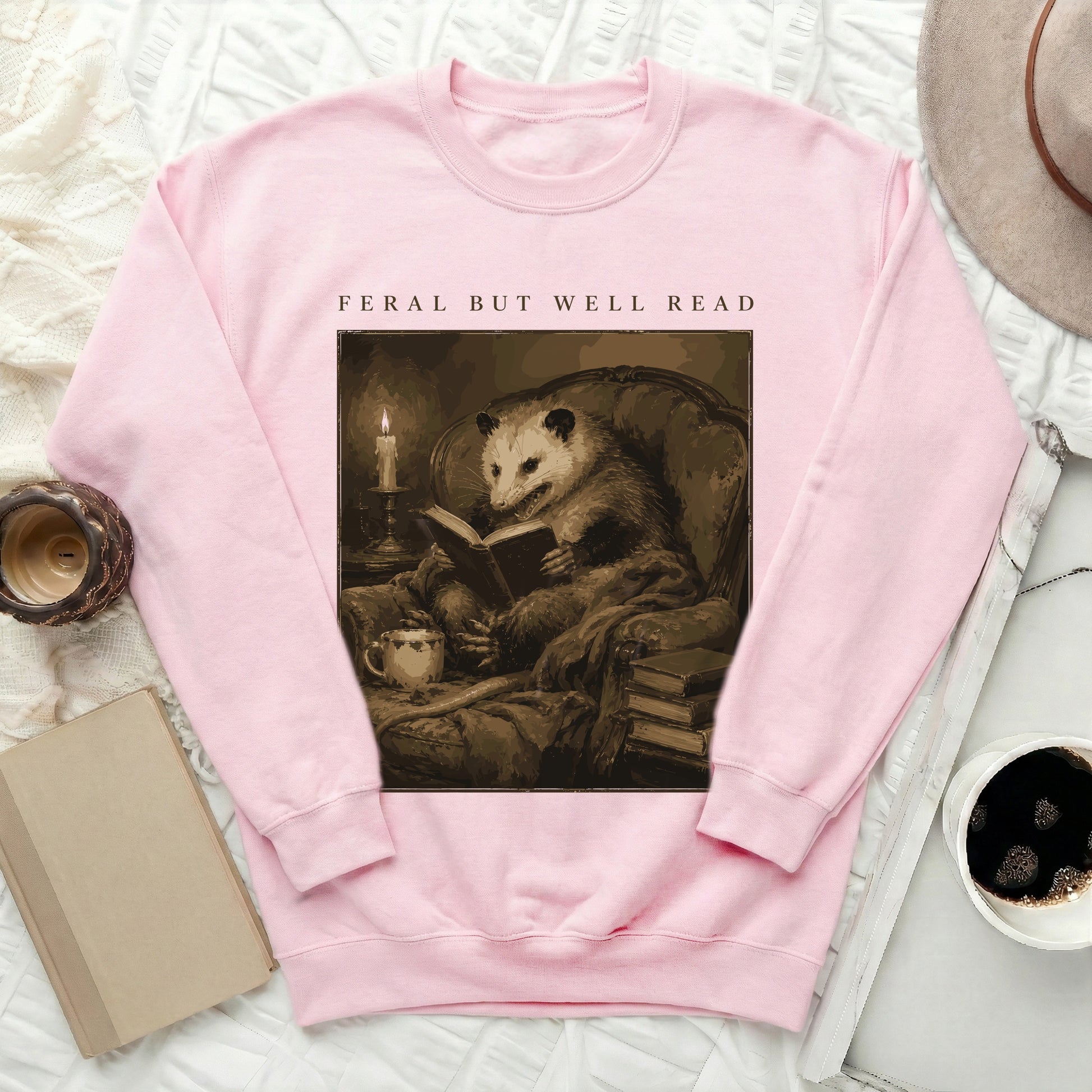 Pink sweatshirt with a graphic of an opossum reading a book and text "FERAL BUT WELL READ".