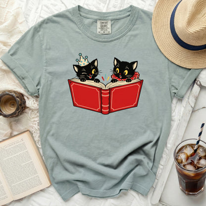 Two black cats read a book, one wearing a party hat and the other a ruff collar.