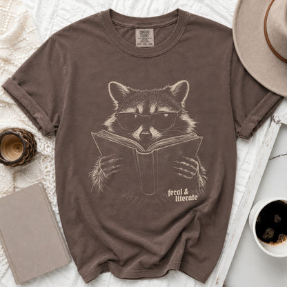 Brown t-shirt with graphic of raccoon wearing glasses reading book, text "feral & literate".