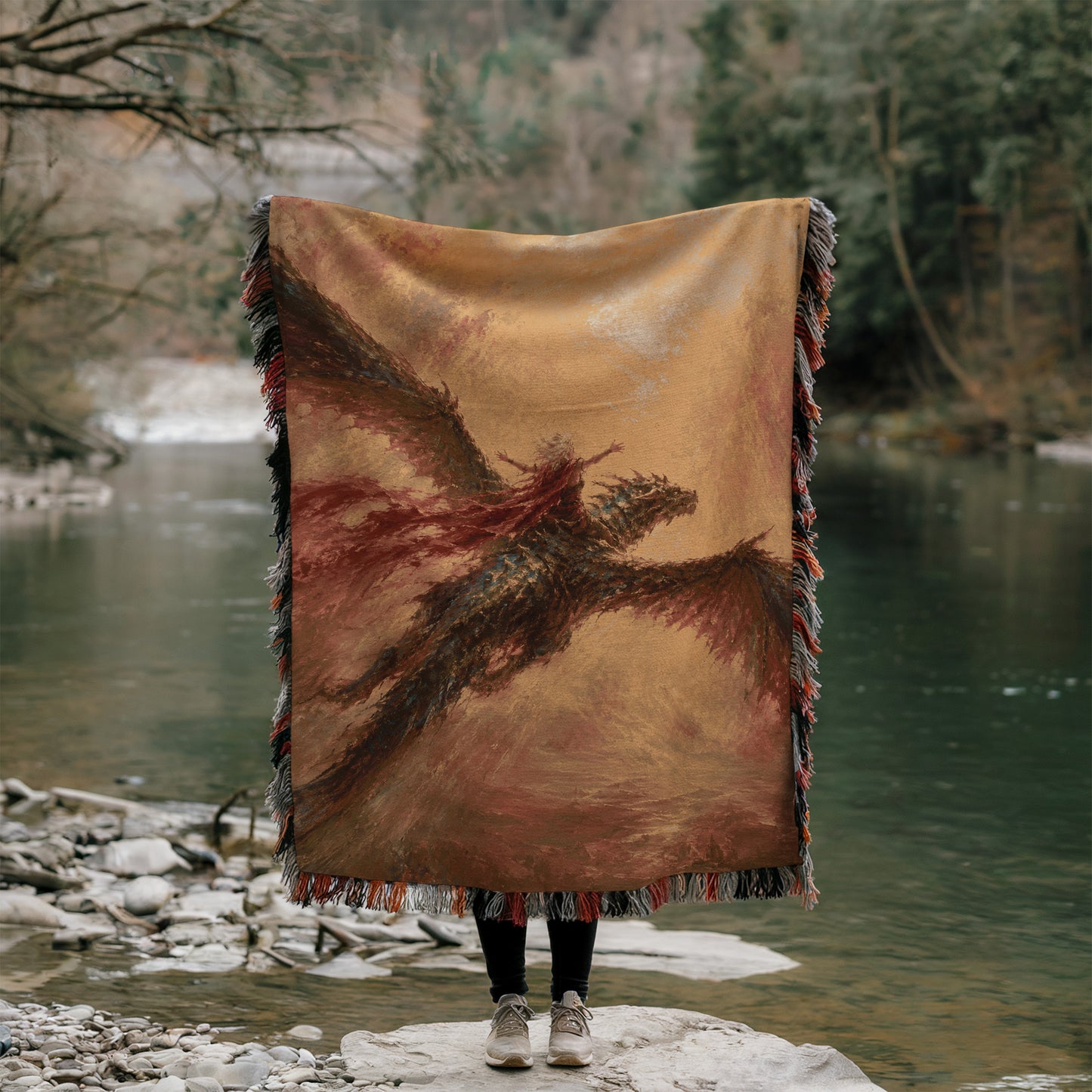 A person stands on a rock, holding a large, colorful blanket that features a dragon design. The person is positioned in front of a body of water, with trees and foliage visible in the background.