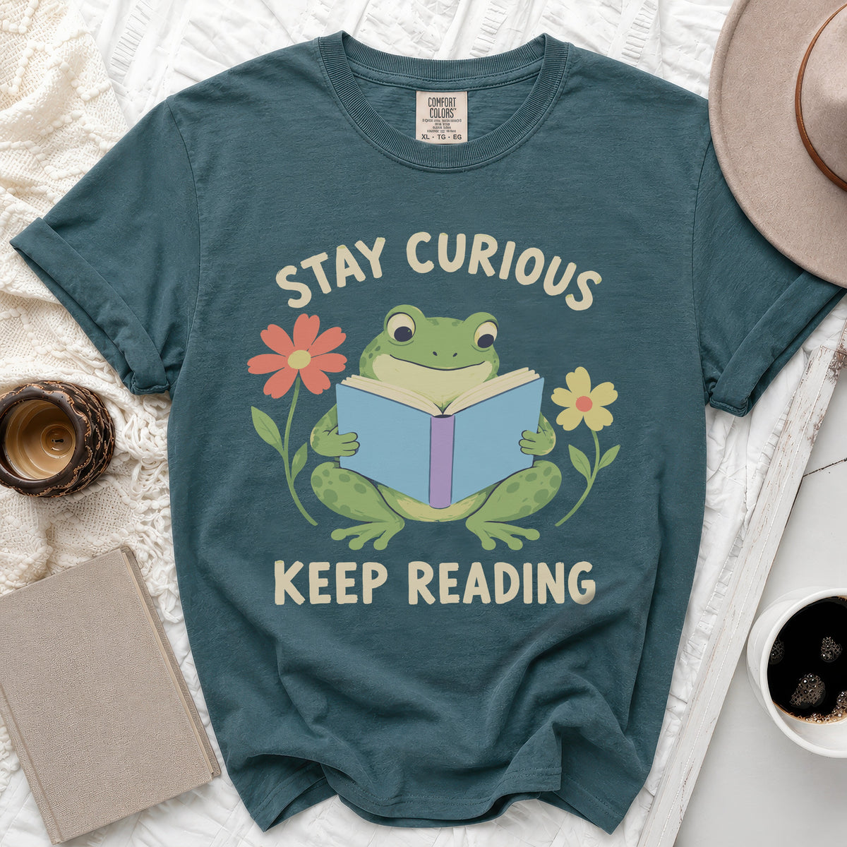 Teal t-shirt with a frog reading a book, surrounded by flowers and text.