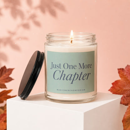 Candle with "Just One More Chapter" text on a shelf with autumn leaves.
