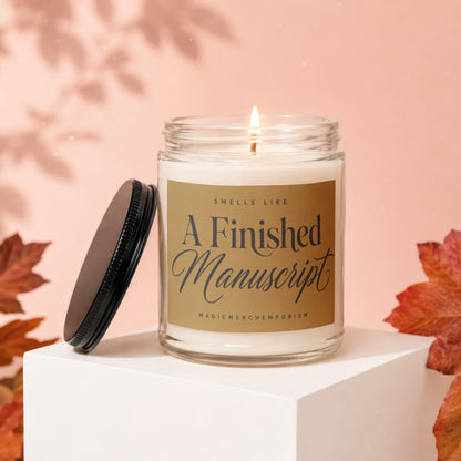 Glass candle with lit flame on golden label reading "Smells Like A Finished Manuscript".