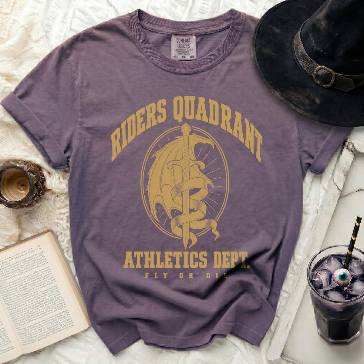 Purple t-shirt with gold Riders Quadrant Athletics Dept dragon and sword graphic, surrounded by books and a drink.