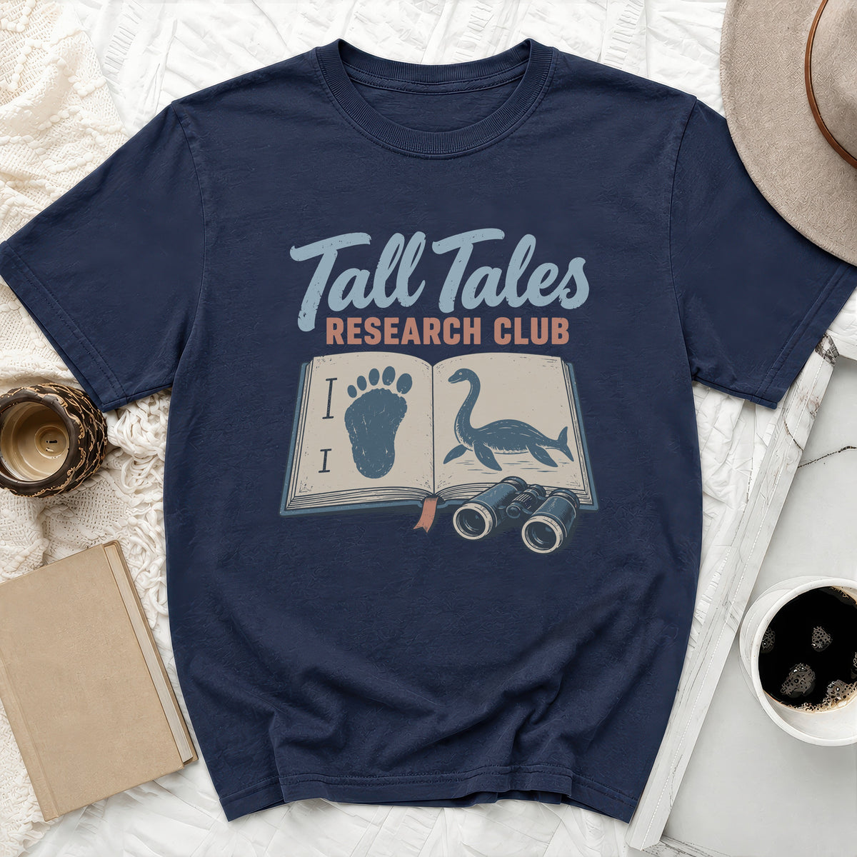 Dark blue t-shirt with "Tall Tales Research Club" text, book, Bigfoot footprint, and Loch Ness Monster.