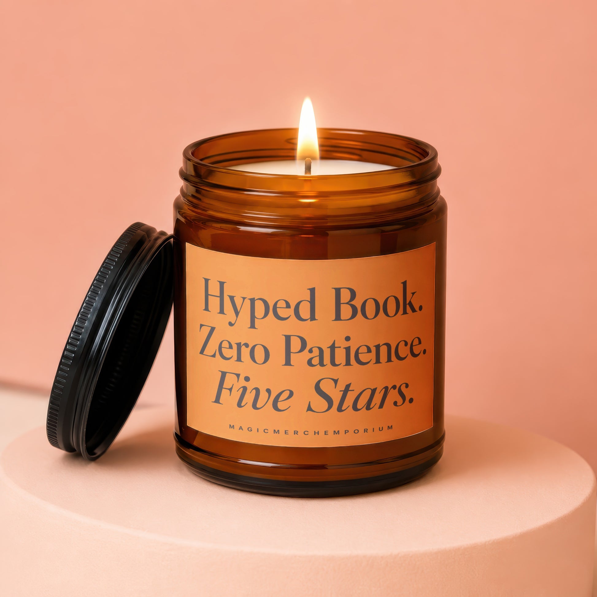 Amber glass candle with lit wick, orange label reads "Hyped Book. Zero Patience. Five Stars."