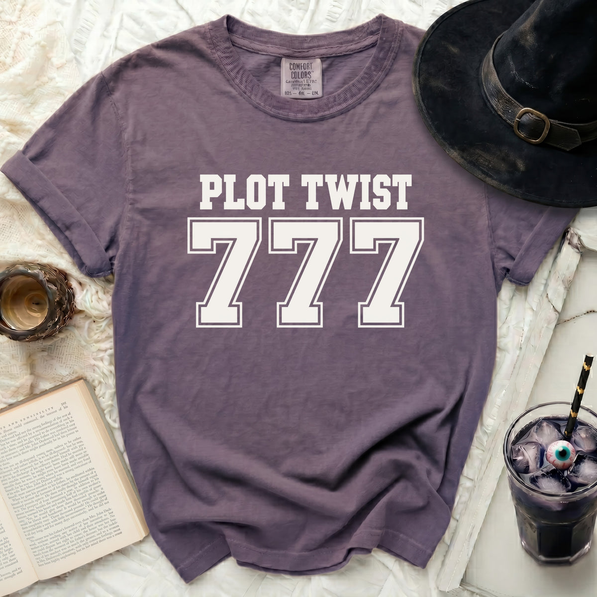 Purple t-shirt with "PLOT TWIST 777" graphic, next to a witch hat and drink.