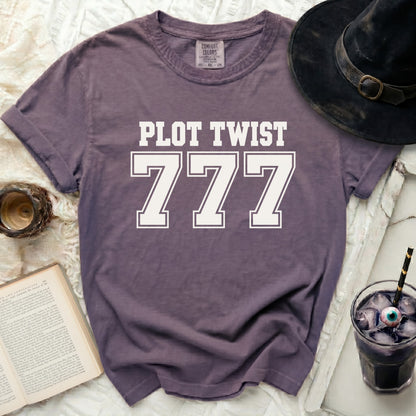 Purple t-shirt with "PLOT TWIST 777" graphic, next to a witch hat and drink.