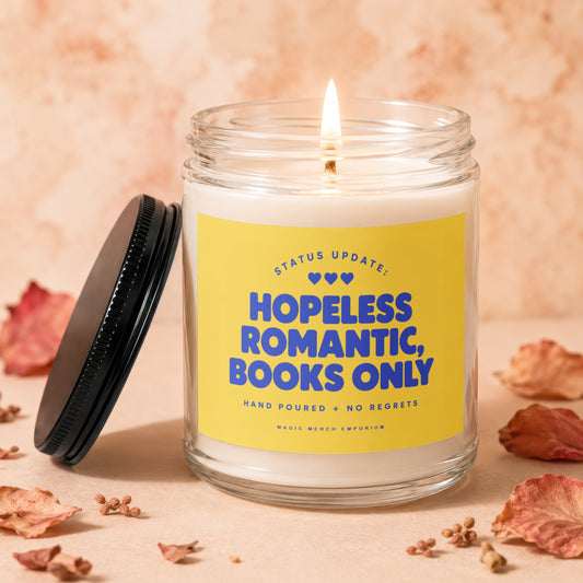 Glass candle with lit wick, yellow label reads "Status Update: Hopeless Romantic, Books Only."