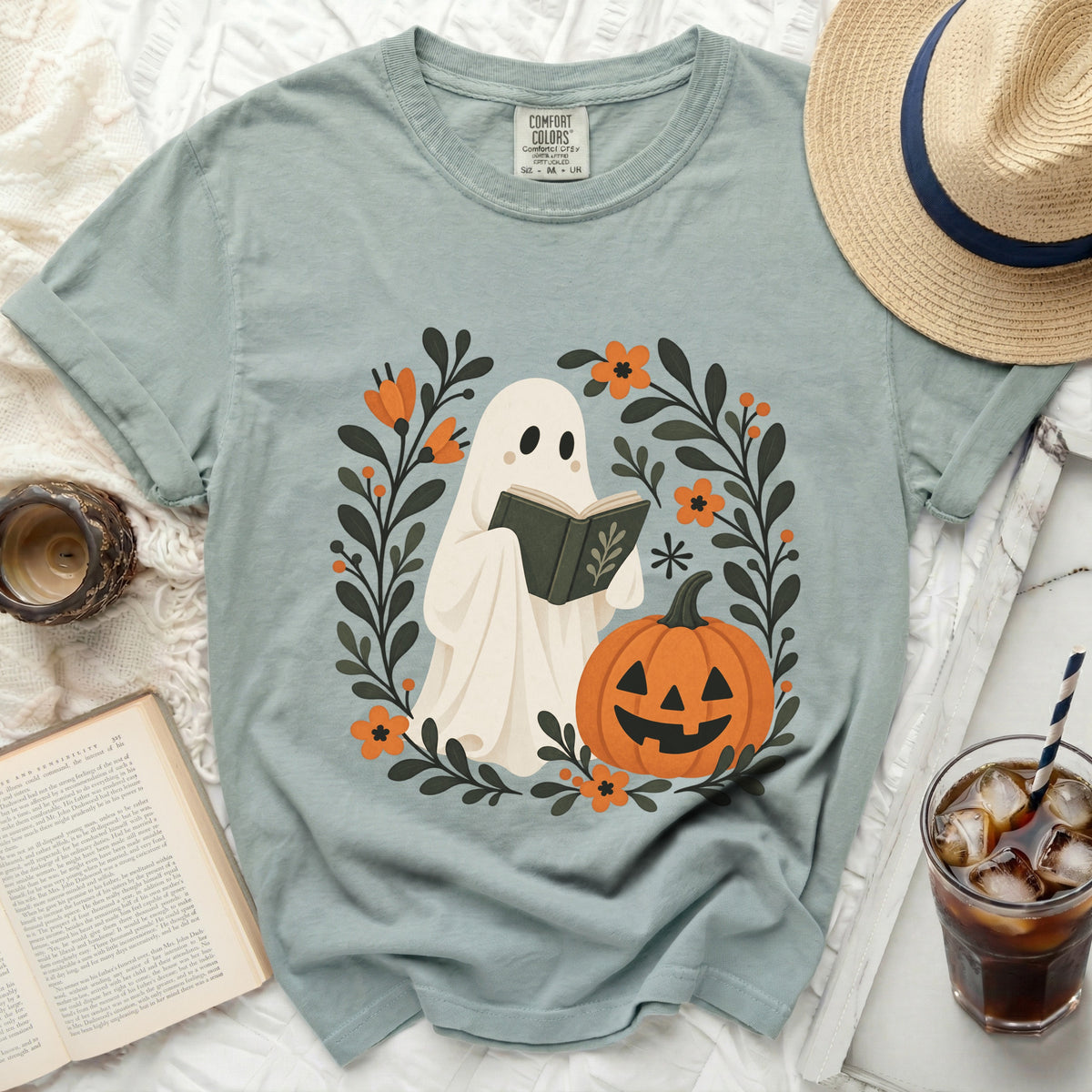 Ghost reading a book surrounded by flowers and a pumpkin graphic on a teal t-shirt.