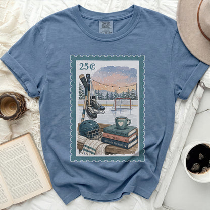 Blue t-shirt with a postage stamp graphic of hockey gear and books.