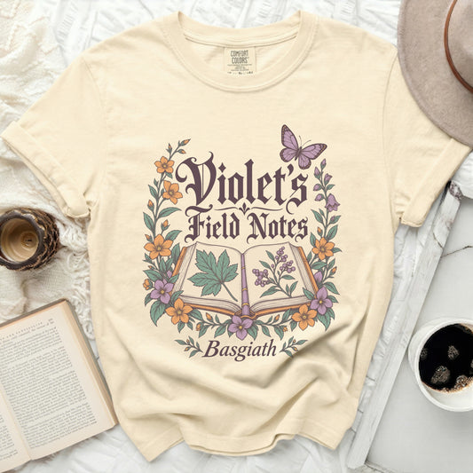 Cream t-shirt with "Violet's Field Notes Basgiath" in gothic font surrounded by flowers and a butterfly.