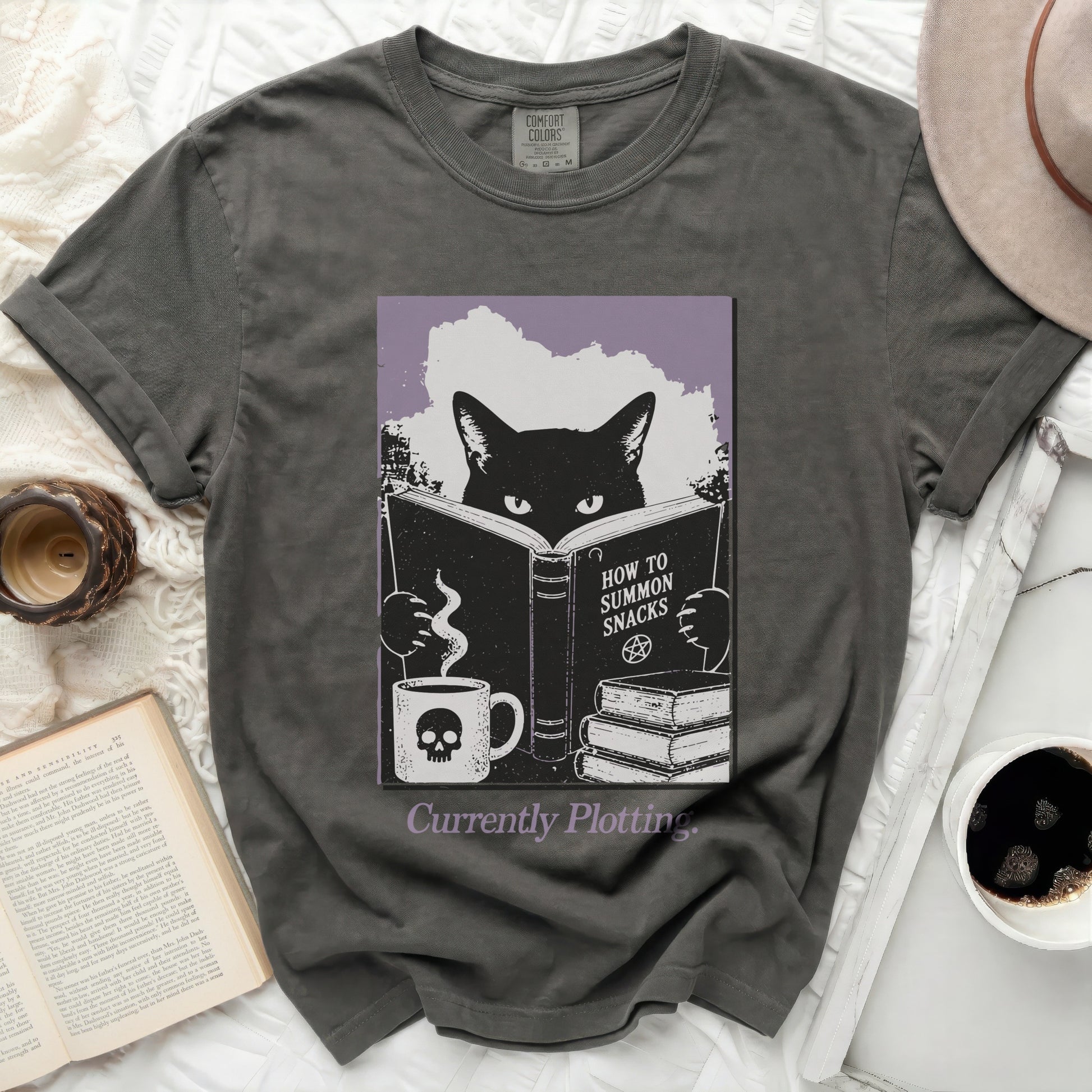 Black cat reading "How to Summon Snacks" with skull mug and books.
