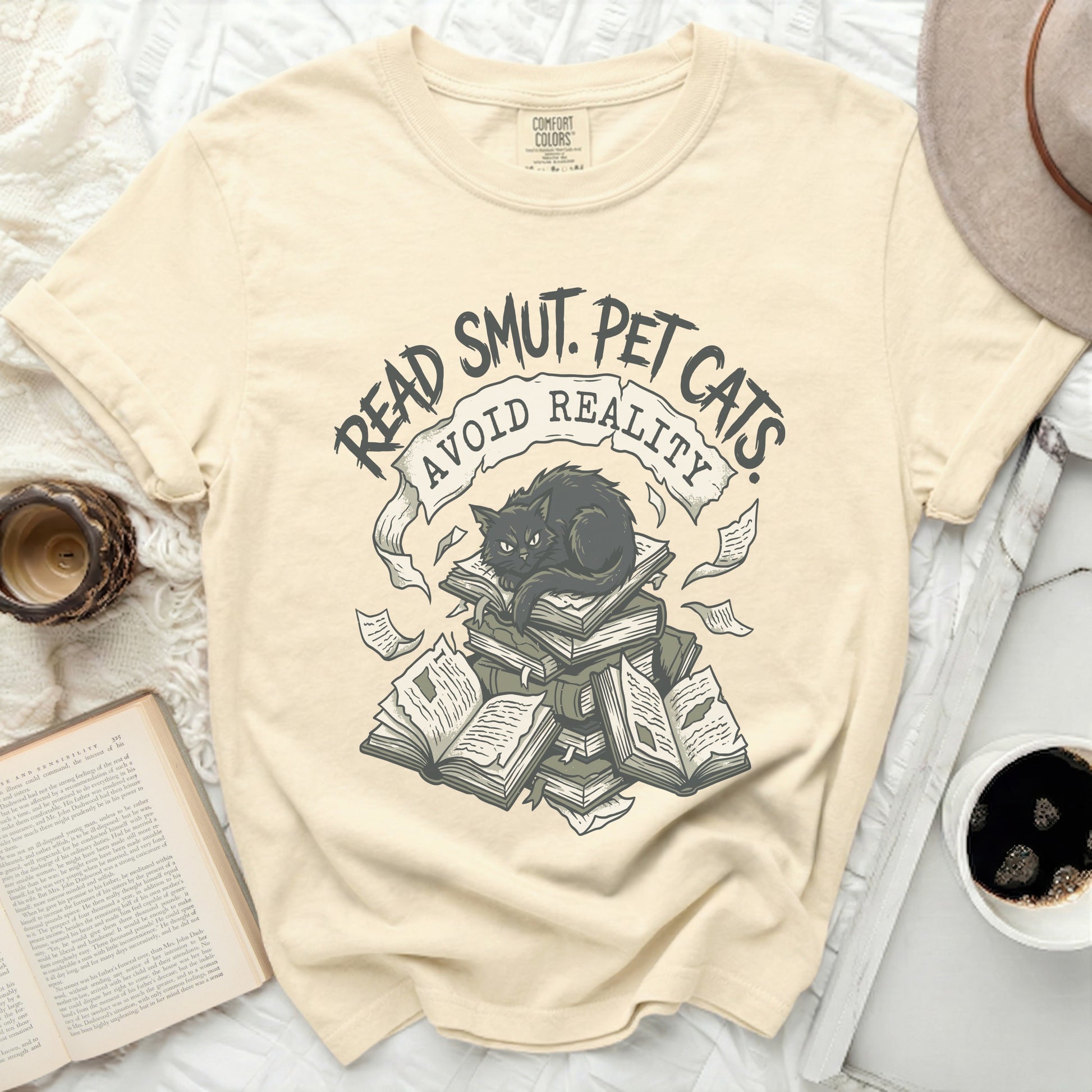 Cream t-shirt with "READ SMUT. PET CATS AVOID REALITY" and a black cat on books.