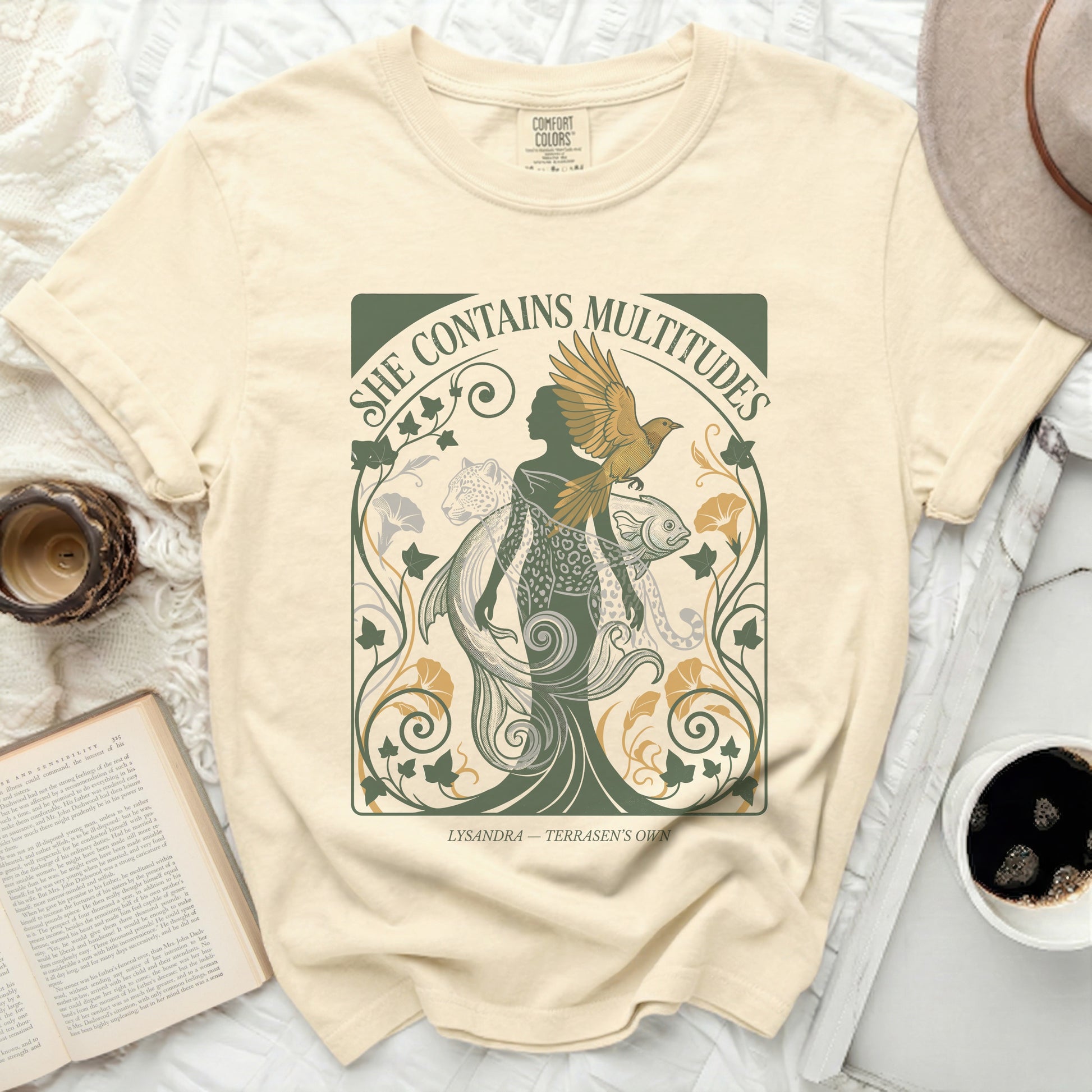 Cream t-shirt with "She Contains Multitudes" text and nature illustration.