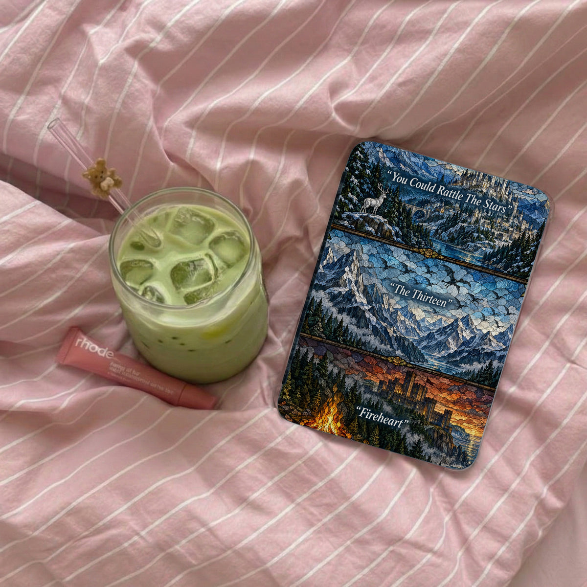 An e-reader in a fantasy-themed stained glass case rests on pink striped bedding next to an iced matcha latte and lip balm.