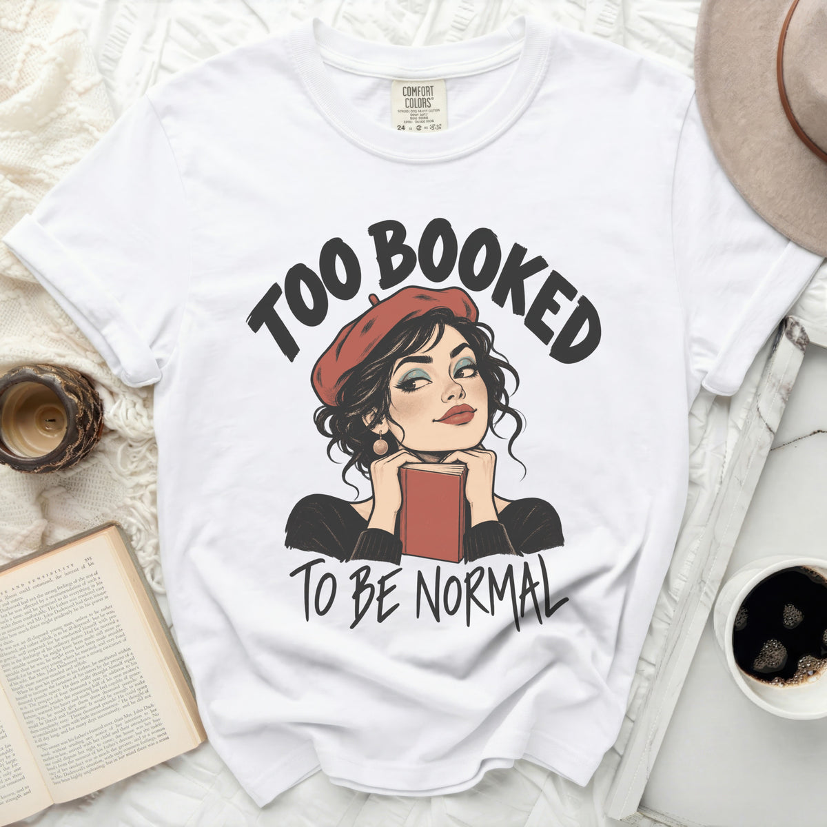 White t-shirt with "TOO BOOKED TO BE NORMAL" text and illustrated woman holding a book.
