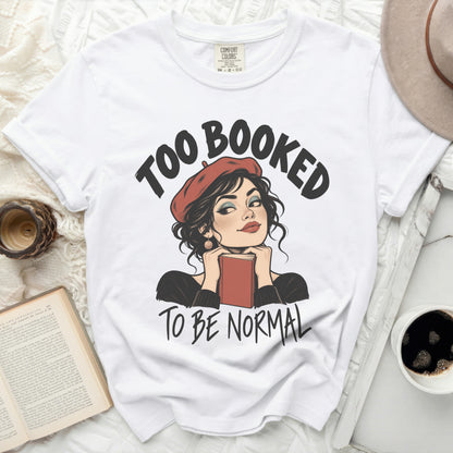 White t-shirt with "TOO BOOKED TO BE NORMAL" text and illustrated woman holding a book.