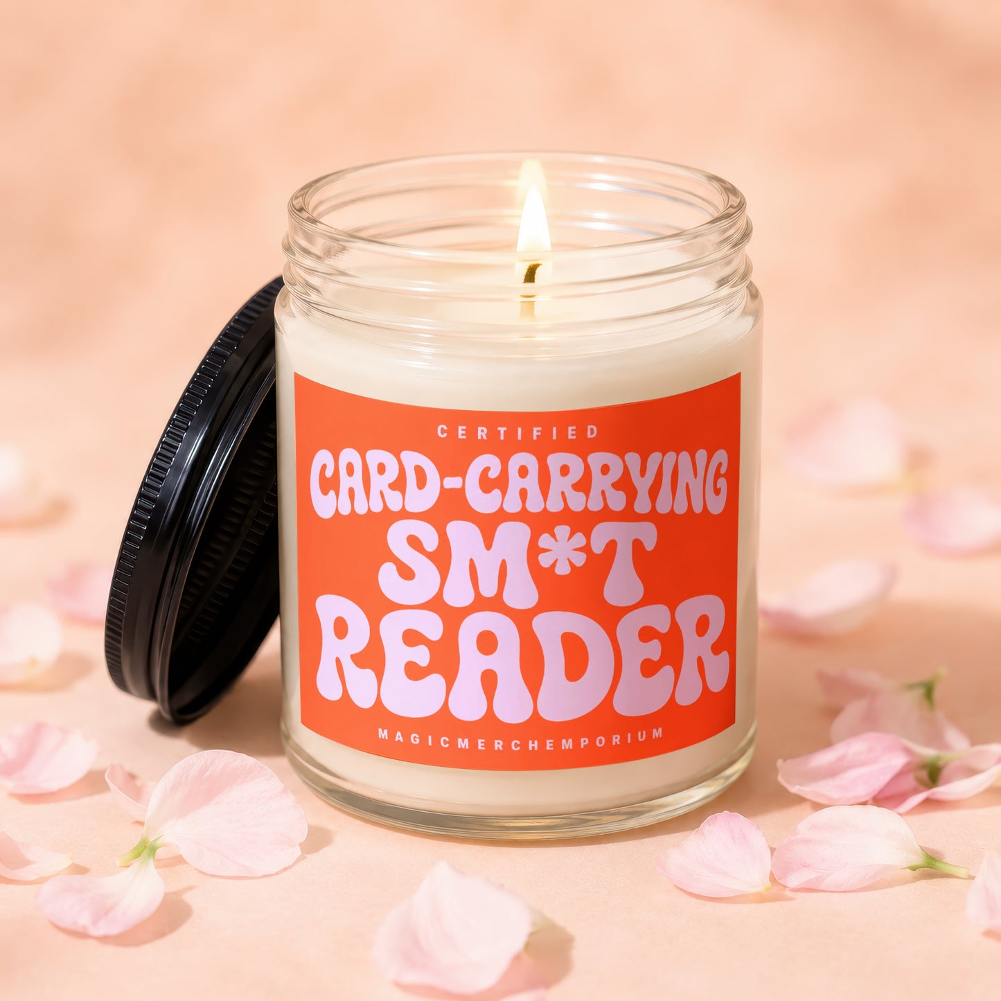Lit candle in glass jar with "Certified Card-Carrying Sm*t Reader" label, pink petals around.