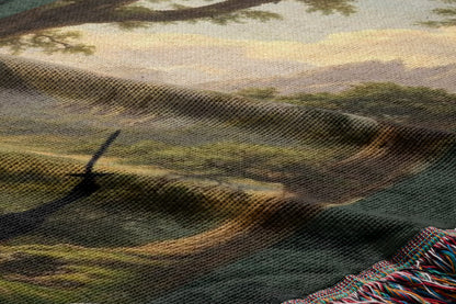 The image is a close-up of a woven fabric with a landscape scene depicted on it. The fabric has a textured appearance, with a mix of green, brown, and gray colors. The scene includes a tree, mountains, and a winding path, creating a serene and natural atmosphere.