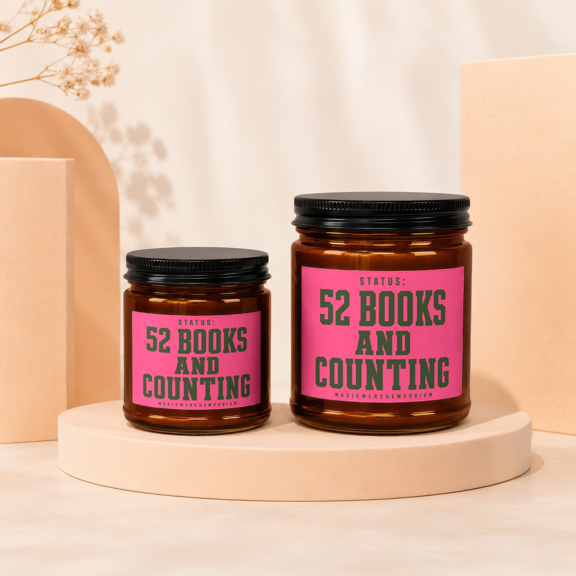 Two amber glass candles with black lids, one smaller than the other, featuring pink labels reading "Status: 52 Books and Counting."