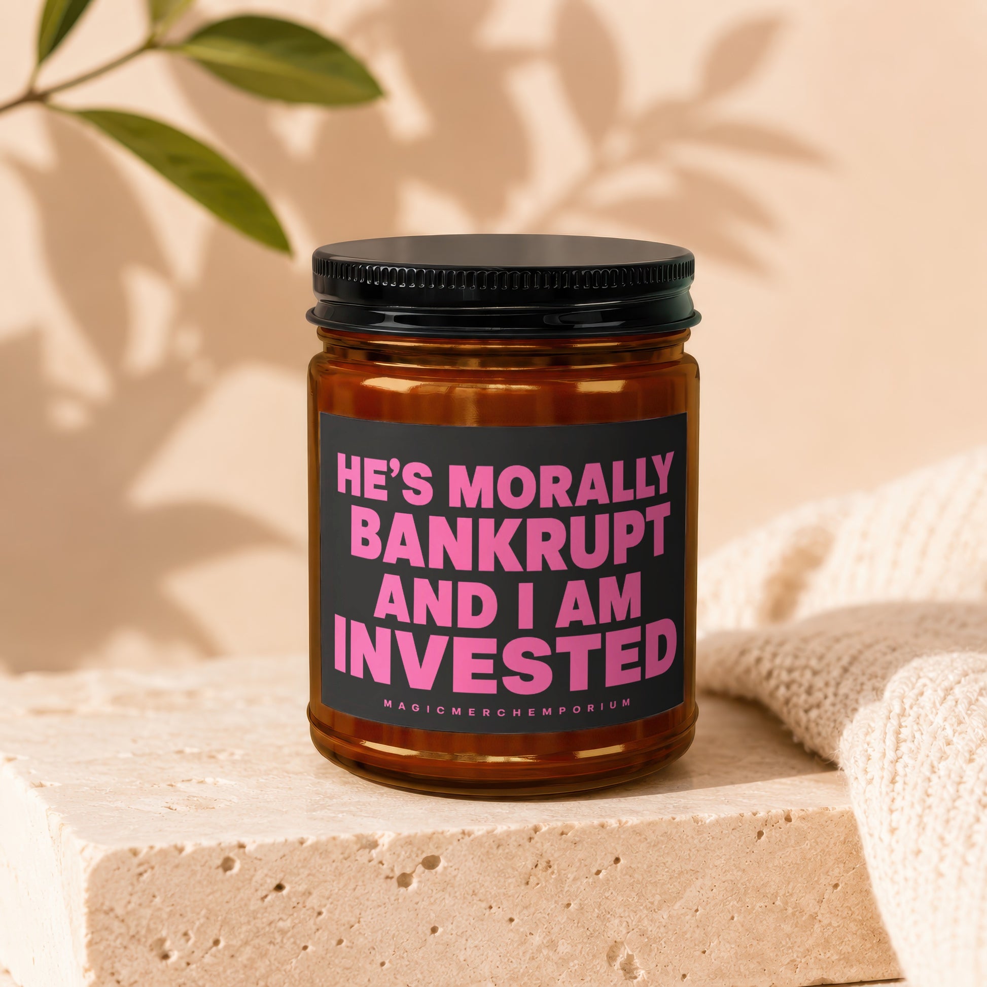 Glass jar candle with black lid and label stating: "HE'S MORALLY BANKRUPT AND I AM INVESTED"
