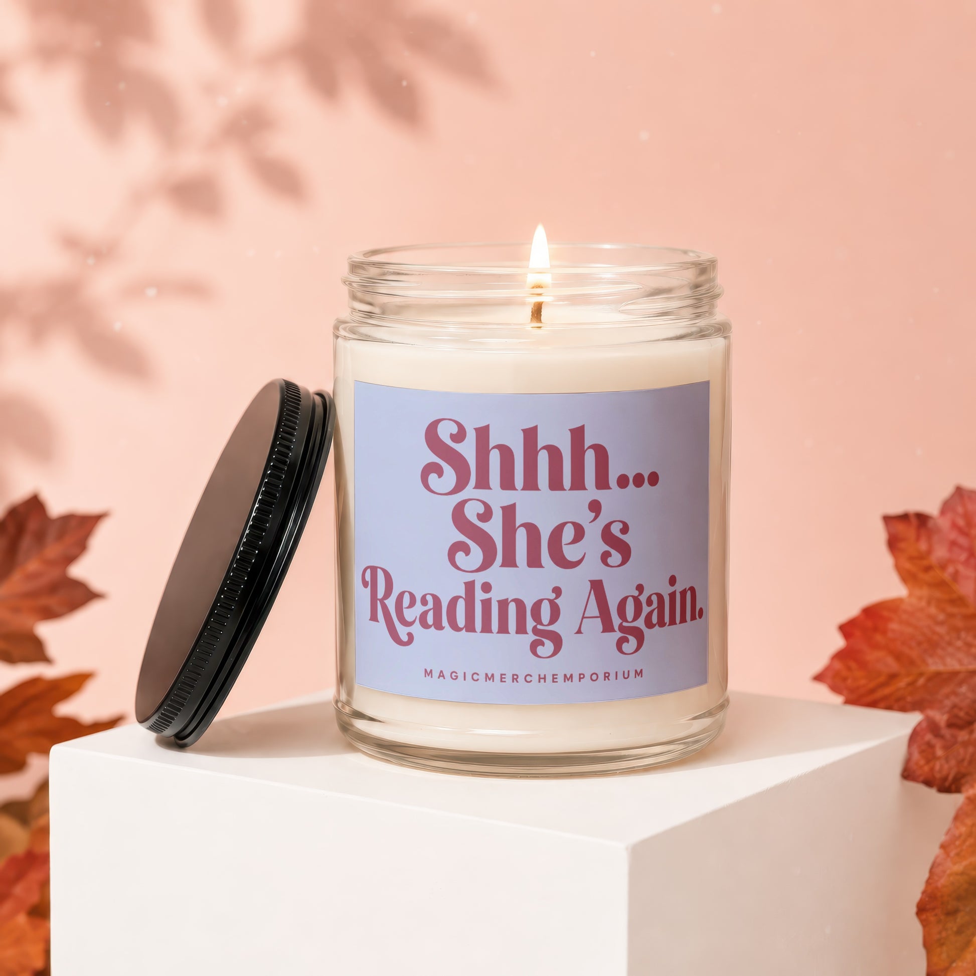 Glass candle with lit wick and lid removed, reads "Shhh... She's Reading Again."