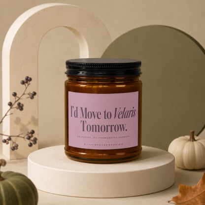 An amber glass candle jar with a pink label reading "I'd Move to Velaris Tomorrow" on a round white platform.