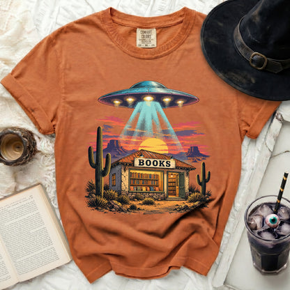 Orange t-shirt with a graphic of a UFO beaming into a desert bookstore.