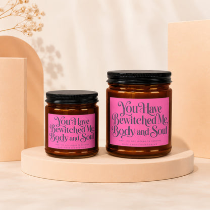 Two amber glass candles with pink labels that say "You Have Bewitched Me, Body and Soul."