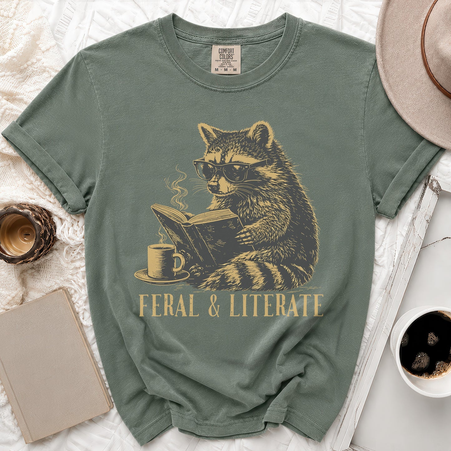 T-shirt with a raccoon wearing glasses reading a book and drinking coffee.