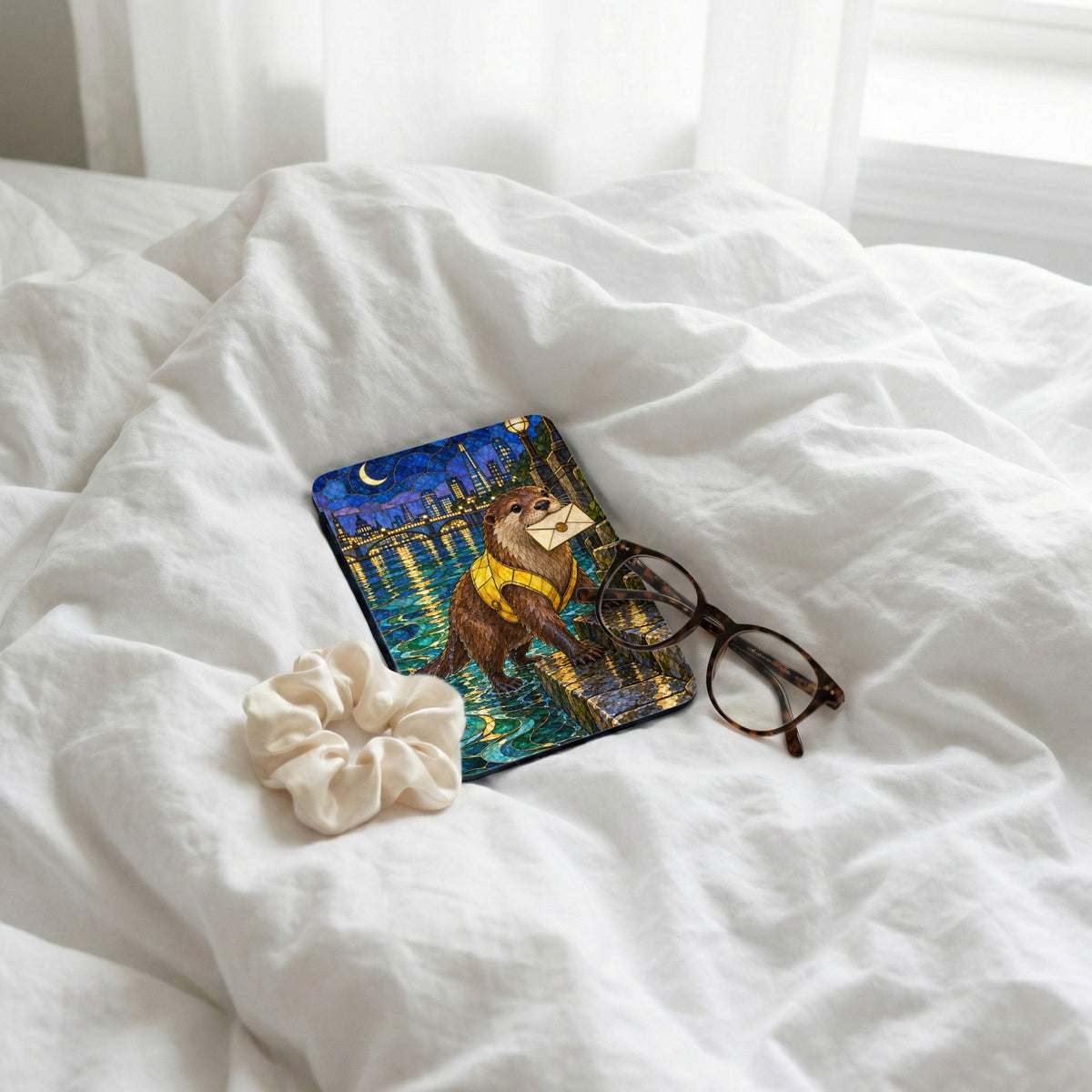 Otter with letter on case, scrunchie, and glasses on white sheets.