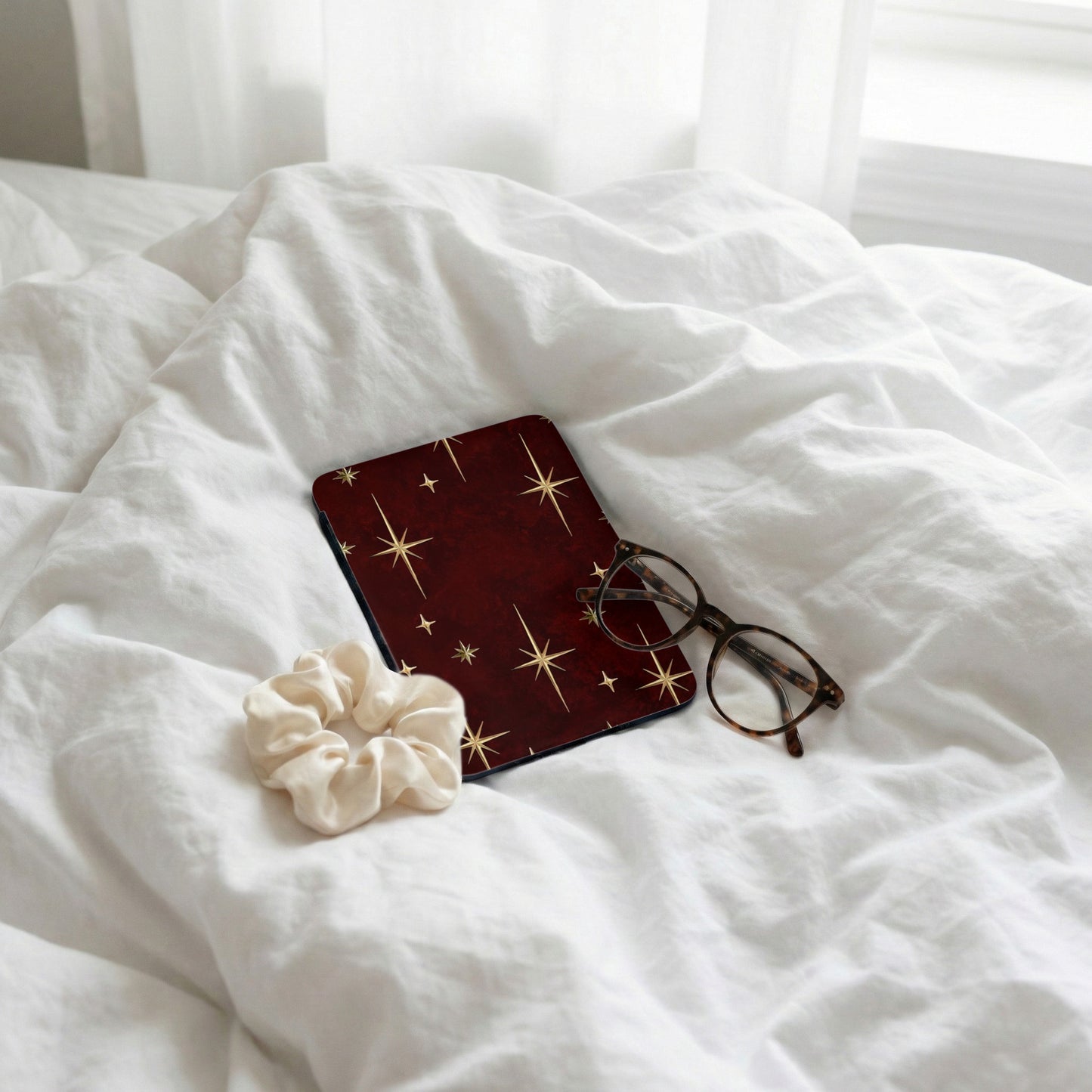 Dark red case with gold starbursts, scrunchie, and glasses on white bedding.