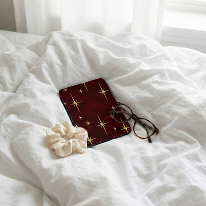 Dark red case with gold starbursts, scrunchie, and glasses on white bedding.