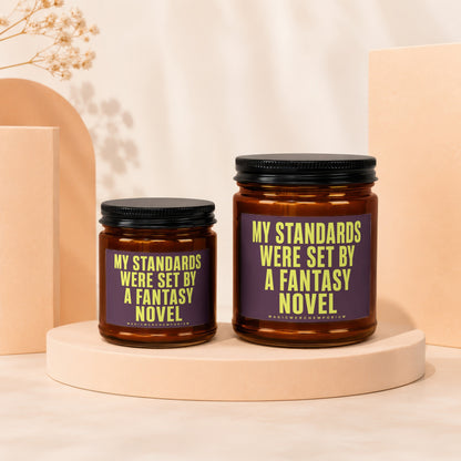 Two amber glass candles with black lids, featuring text "My Standards Were Set By A Fantasy Novel".