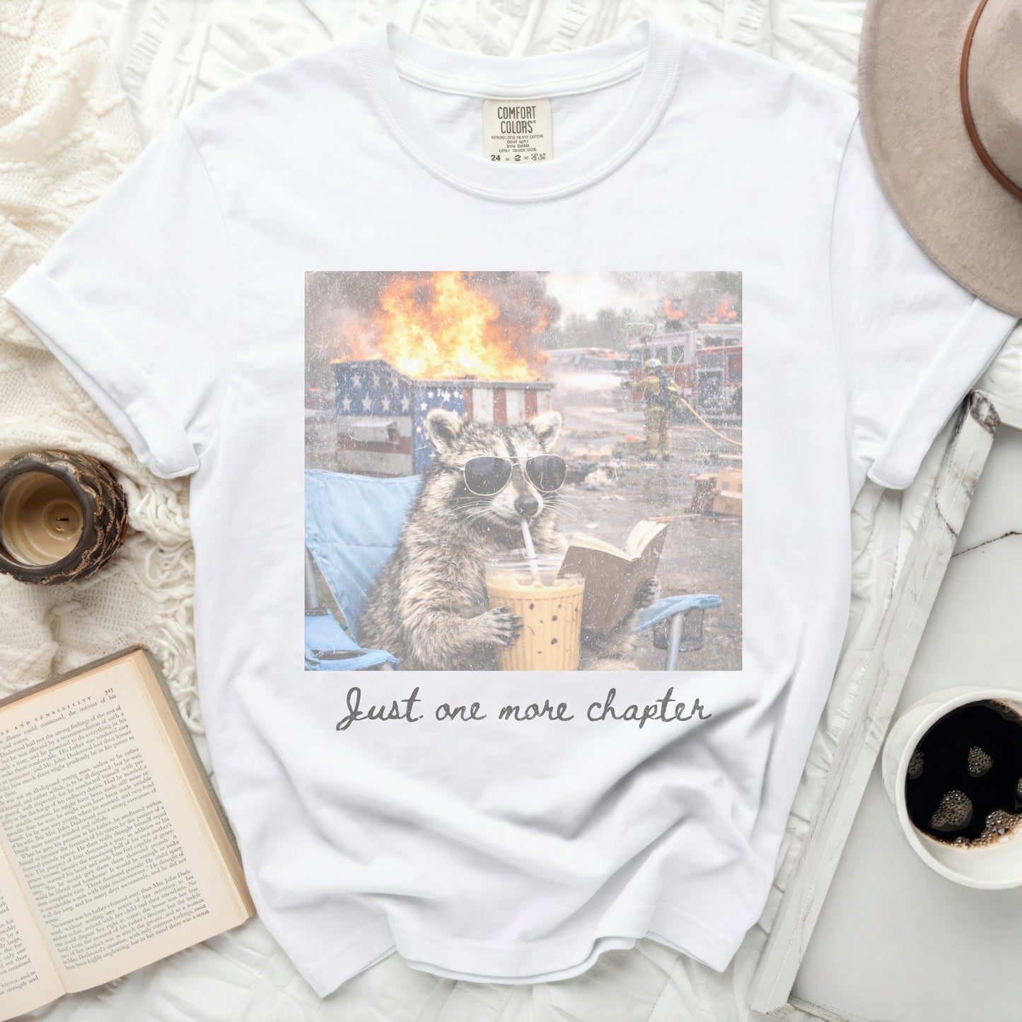 White Comfort Colors t-shirt with raccoon in sunglasses reading and drinking.