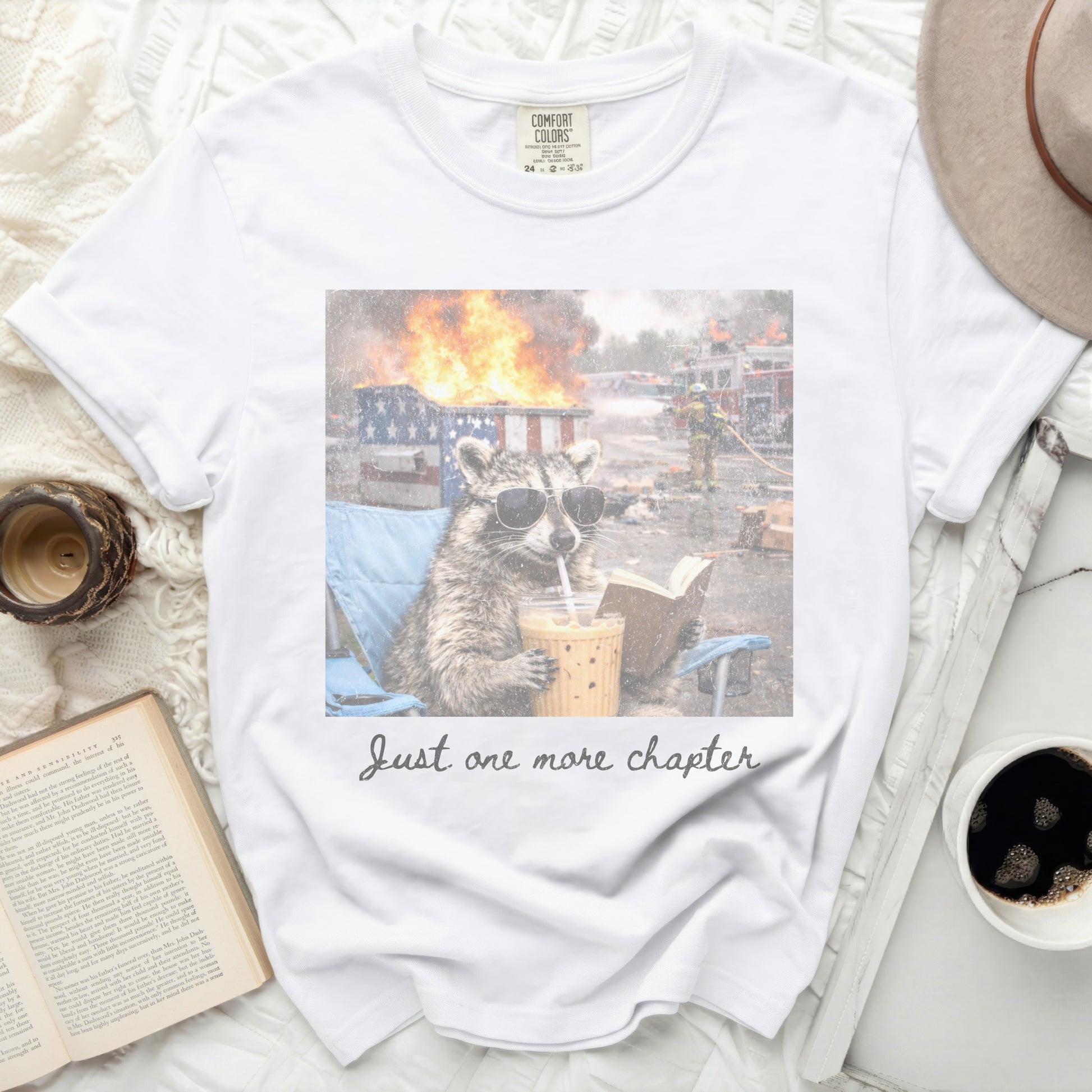 White Comfort Colors t-shirt with raccoon in sunglasses reading and drinking.