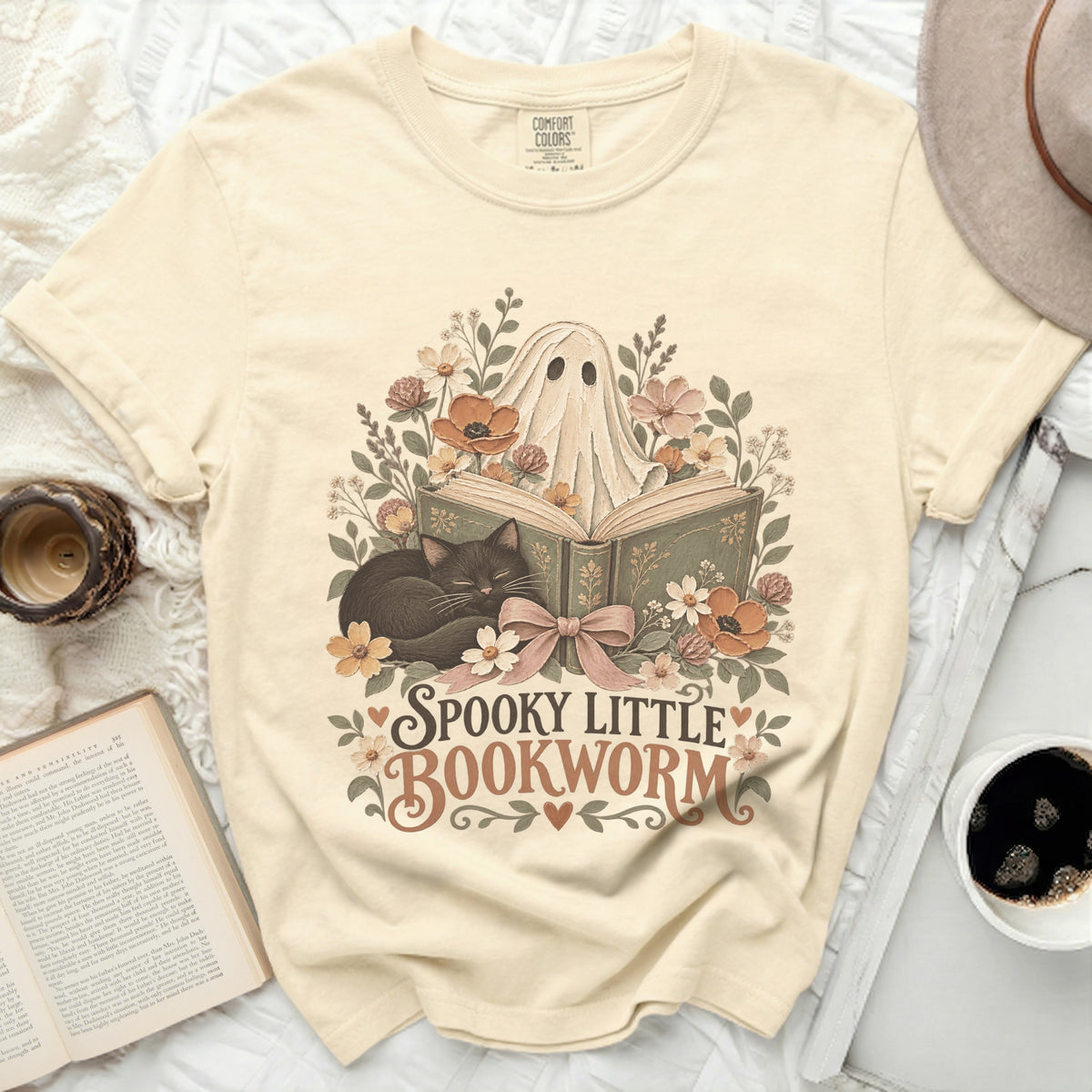 A cream t-shirt with a ghost reading a book, a black cat, and flowers, reads "Spooky Little Bookworm".