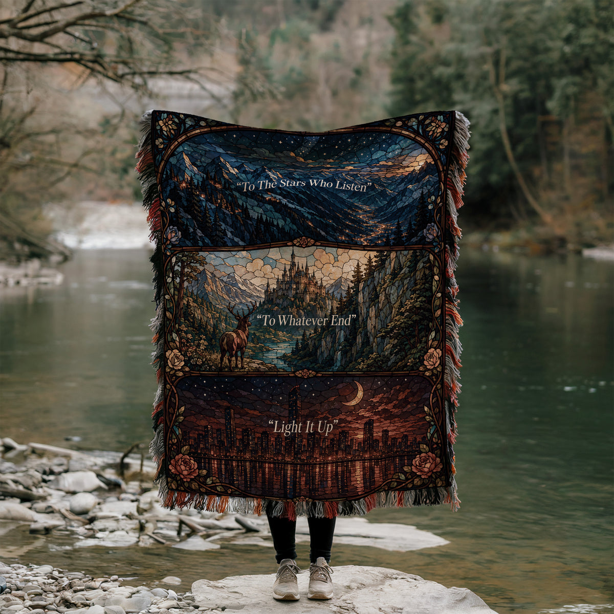 A person stands on a rock, holding a colorful blanket with a landscape scene and text printed on it, in front of a river and forest.