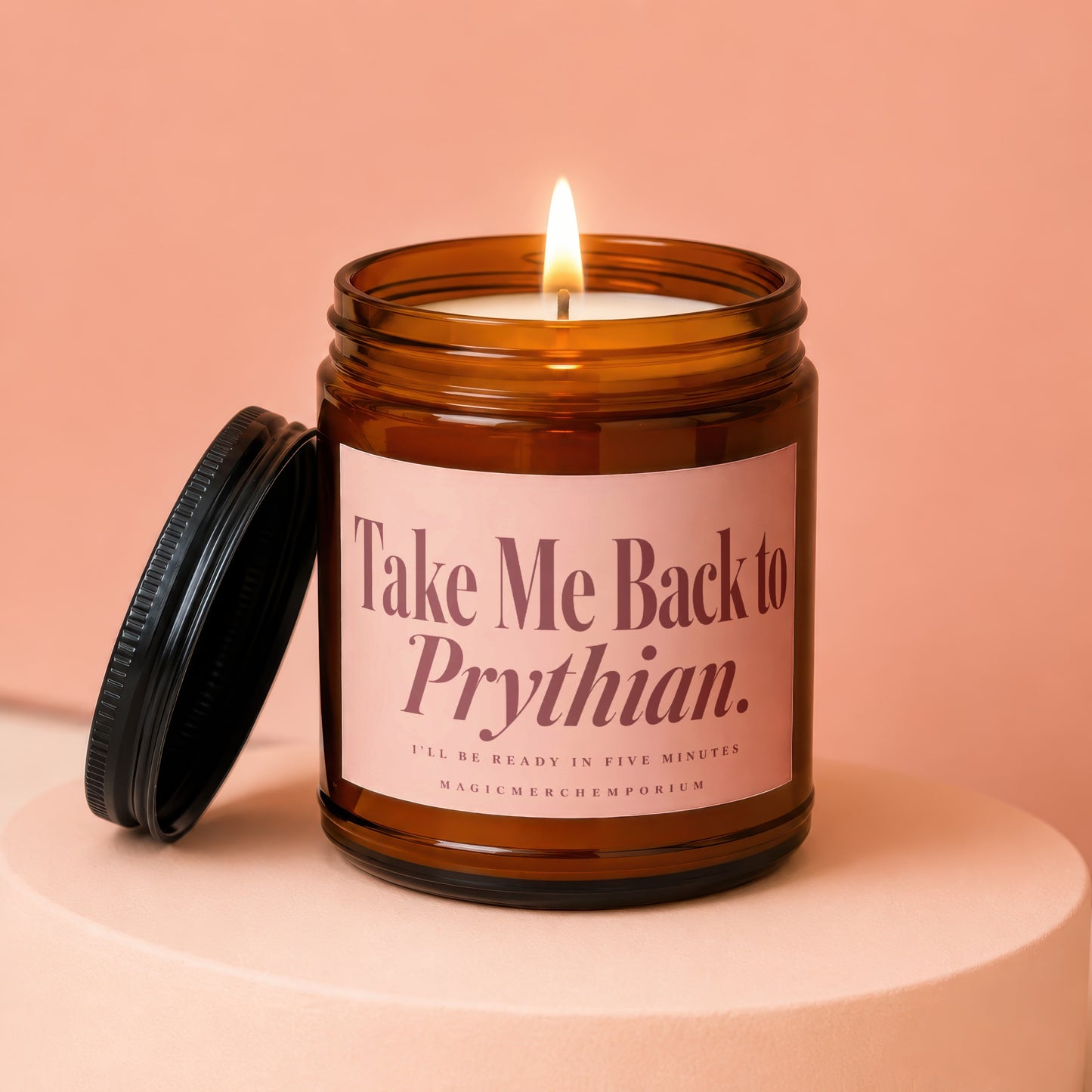 A lit amber glass jar candle with a pink label reading Take Me Back to Prythian sits on a pink pedestal with its black lid resting beside it against a matching pink background.