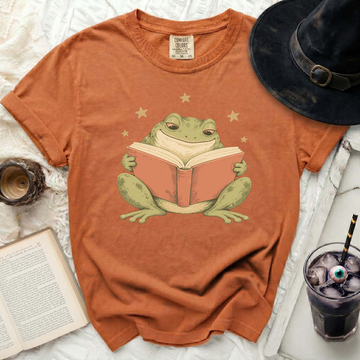 Orange t-shirt with a graphic of a smiling frog reading a book and stars.
